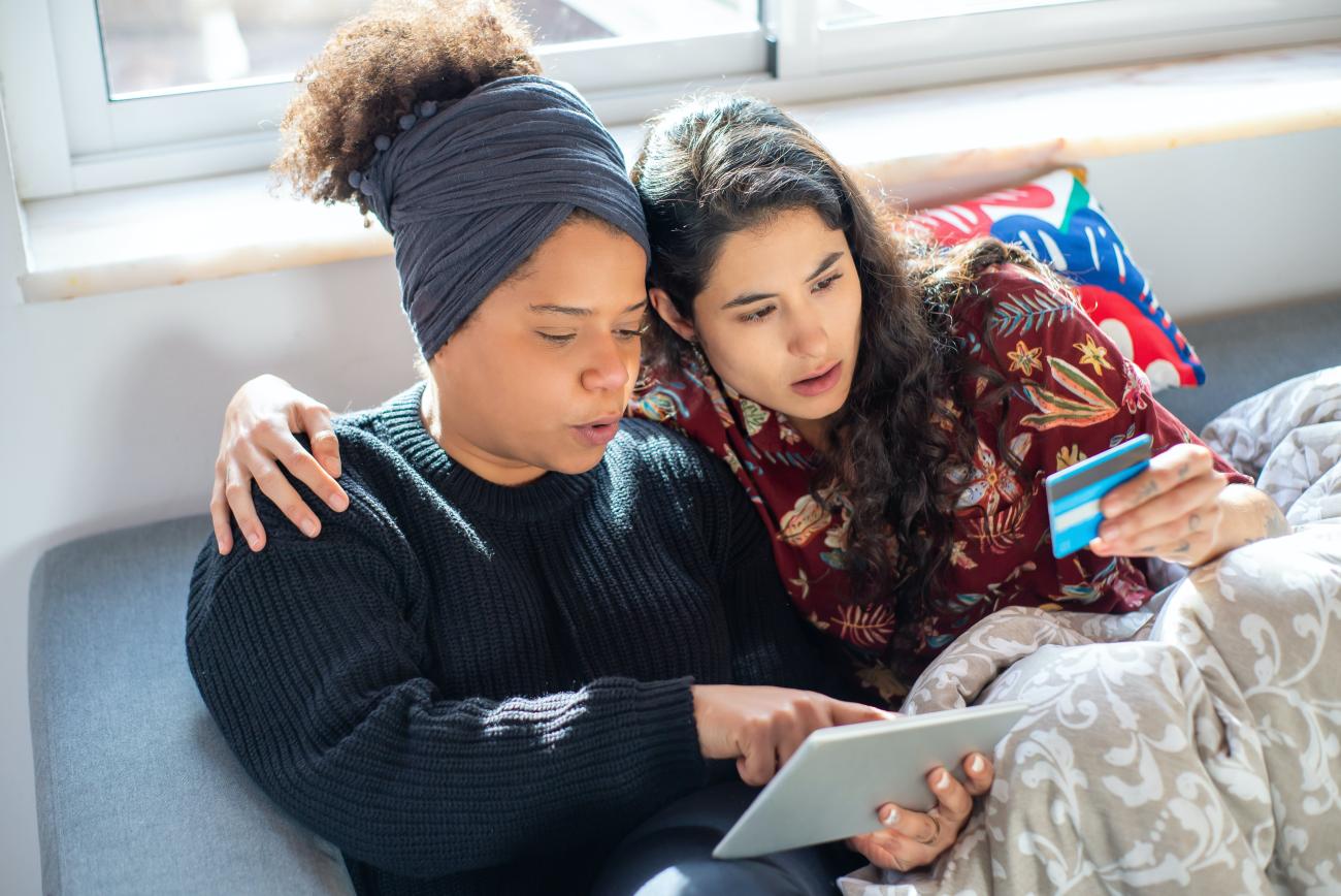 Two women of color sit on a couch with a blanket over their legs. The woman on the left has her natural hair up in a wrap, the other woman sits to the first woman's left with her arm around her. The women are looking at a credit card and tablet.