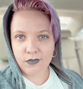 A headshot of a white woman looking straight into the camera with short pink hair and grey lipstick, a nose hoop, and a grey hoodie with the hood pulled up.