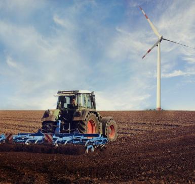 Image: tractor on a field with a wind turbine. Topic: Regenerative Agriculture.