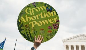A white hand holding a round, green sign that reads: "Grow Abortion Power" in front of the Supreme Court building