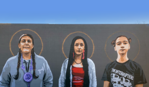 Mural painted on the outside of Native American Bank. There are three women of different ages standing side by side with halos around their heads to represent what Native American Bank honors and celebrates.