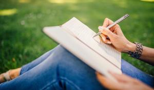 A woman's hand writes in a notebook resting on her knees. She's sitting in a grass field. Sustainable creative accessories.