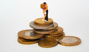 Two clay figures in a romantic embrace stand atop a pile of gold coins. Valentine's Day love money.