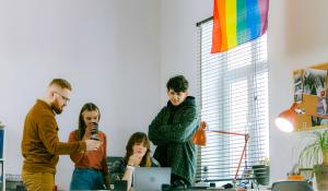 Four white people at a desk. A woman sits in the middle in front of a laptop, the other three stand around her, looking at the laptop and talking. There's a rainbow flag on the wall beside them. LGBTQ-owned