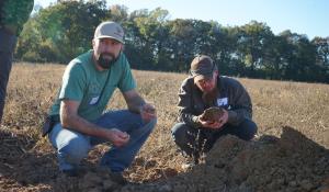two men squatting in the dirt of a field, holding clumps of soil in their hands and examining them.