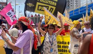 A woman in a patterened white dress stands amidst climate protesters with her arms raised victoriously.