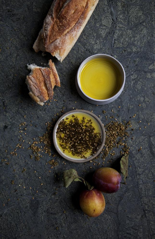A vertical image, showing a baguette, a small white jar of Canaan Palestine olive oil and Za'atar, as well as plums, on black marble.
