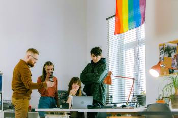 Four white people at a desk. A woman sits in the middle in front of a laptop, the other three stand around her, looking at the laptop and talking. There's a rainbow flag on the wall beside them. LGBTQ-owned